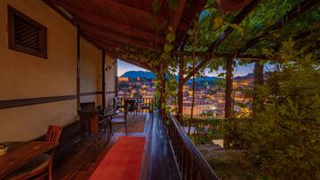 Um deck de madeira com carpete vermelho, cadeiras de madeira e uma mesa, com vista para um horizonte urbano com edifícios e uma montanha ao fundo.