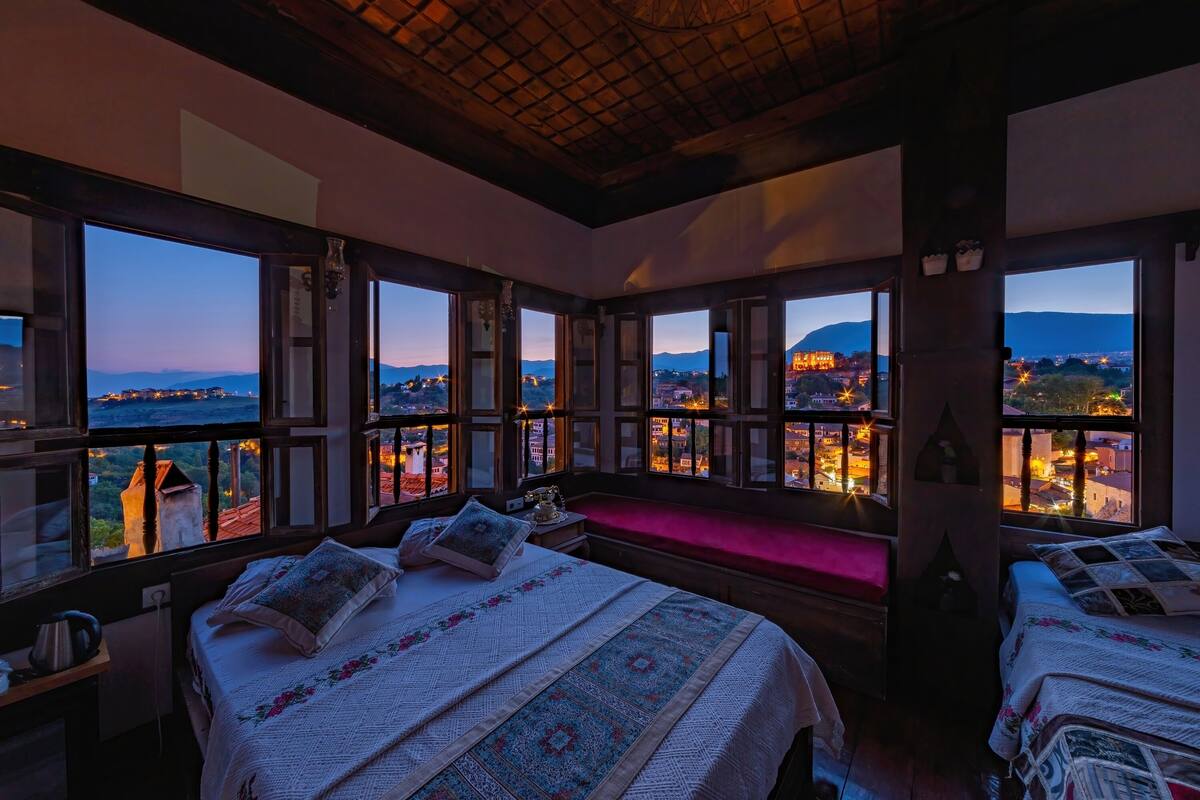 Un dormitorio con ventanas amplias, una cama con colcha estampada, una mesita de noche y un banco.