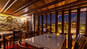 Un restaurante con pared de piedra, techo de madera y grandes ventanales con vista a un paisaje urbano nocturno.