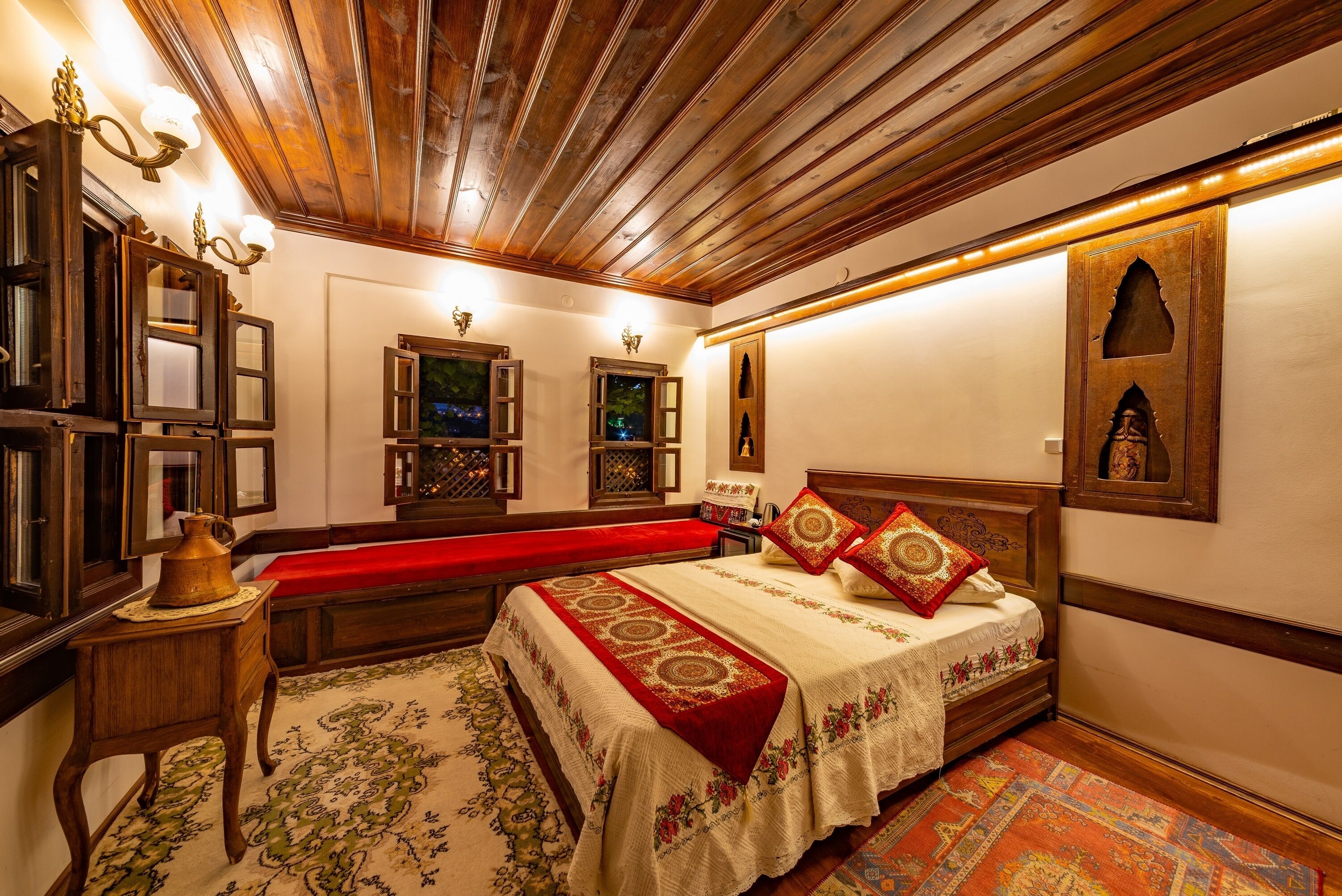 A traditional room with a wooden ceiling, a large bed with red and white bedding, a bench, and a small table with a lamp.