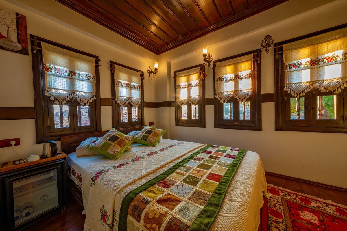Un dormitorio con una cama grande, varias ventanas con cortinas de encaje, techo de madera y una alfombra roja con estampado.
