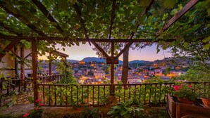 Un jardín con una pérgola, plantas en macetas y vistas a un paisaje urbano al atardecer.