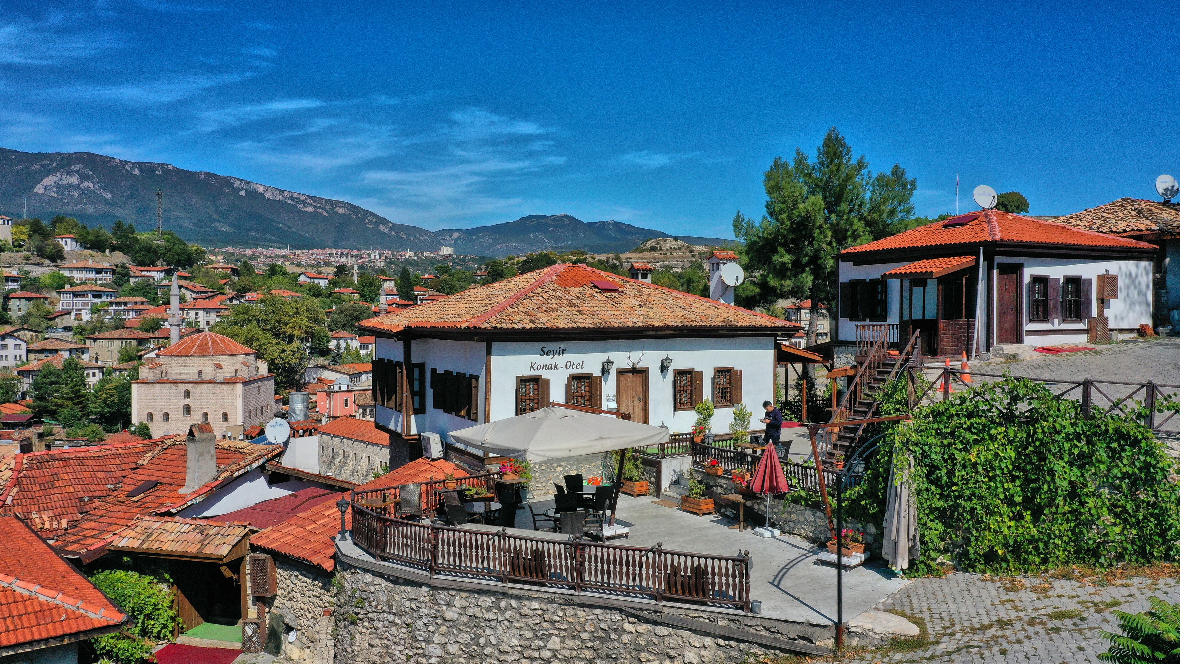 A traditional building with a tiled roof, a balcony with outdoor seating, and a view of a town with mountains in the background.