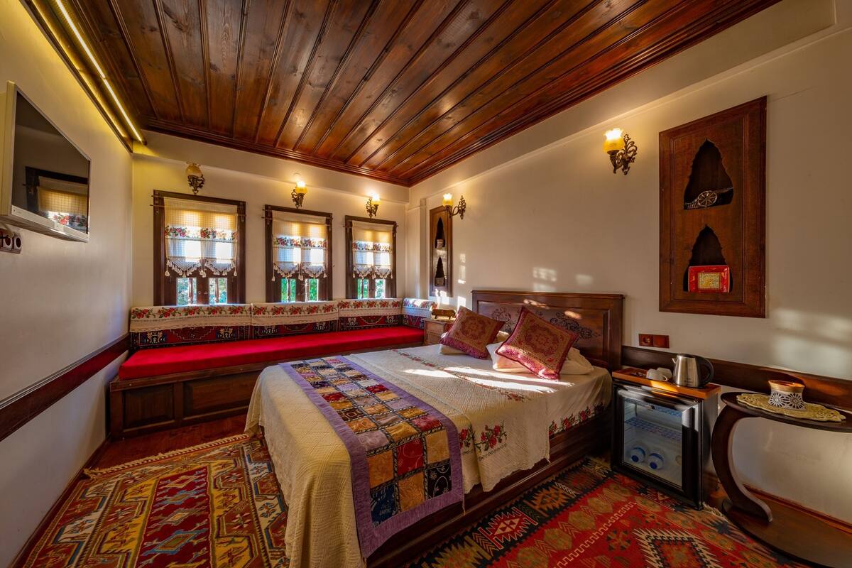 Una habitación tradicional con techo de madera, una cama grande con colcha, un banco y una pequeña mesa con hervidor.