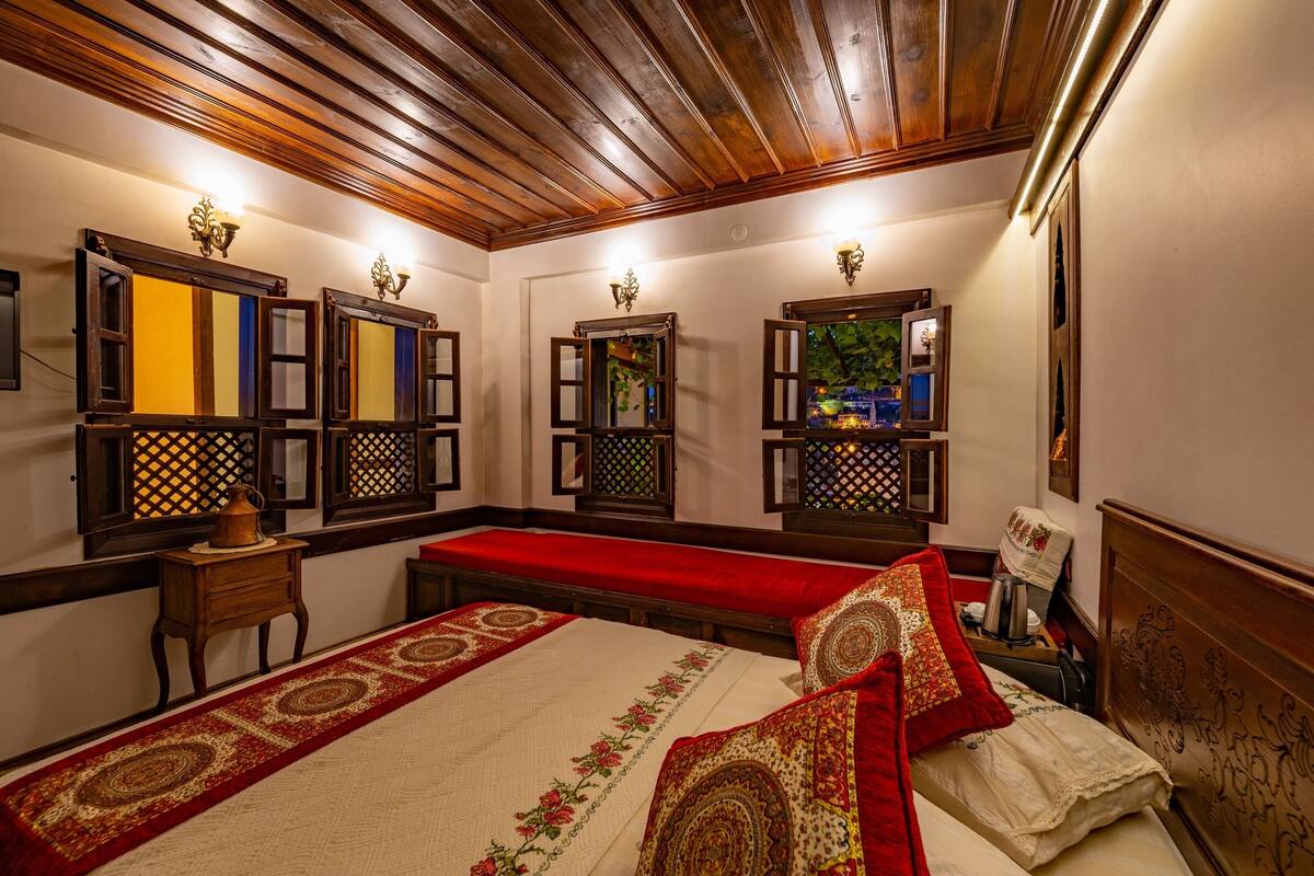 Una habitación tradicional con techo de madera, una cama grande con ropa de cama roja y blanca, y varias ventanas con marcos decorativos.