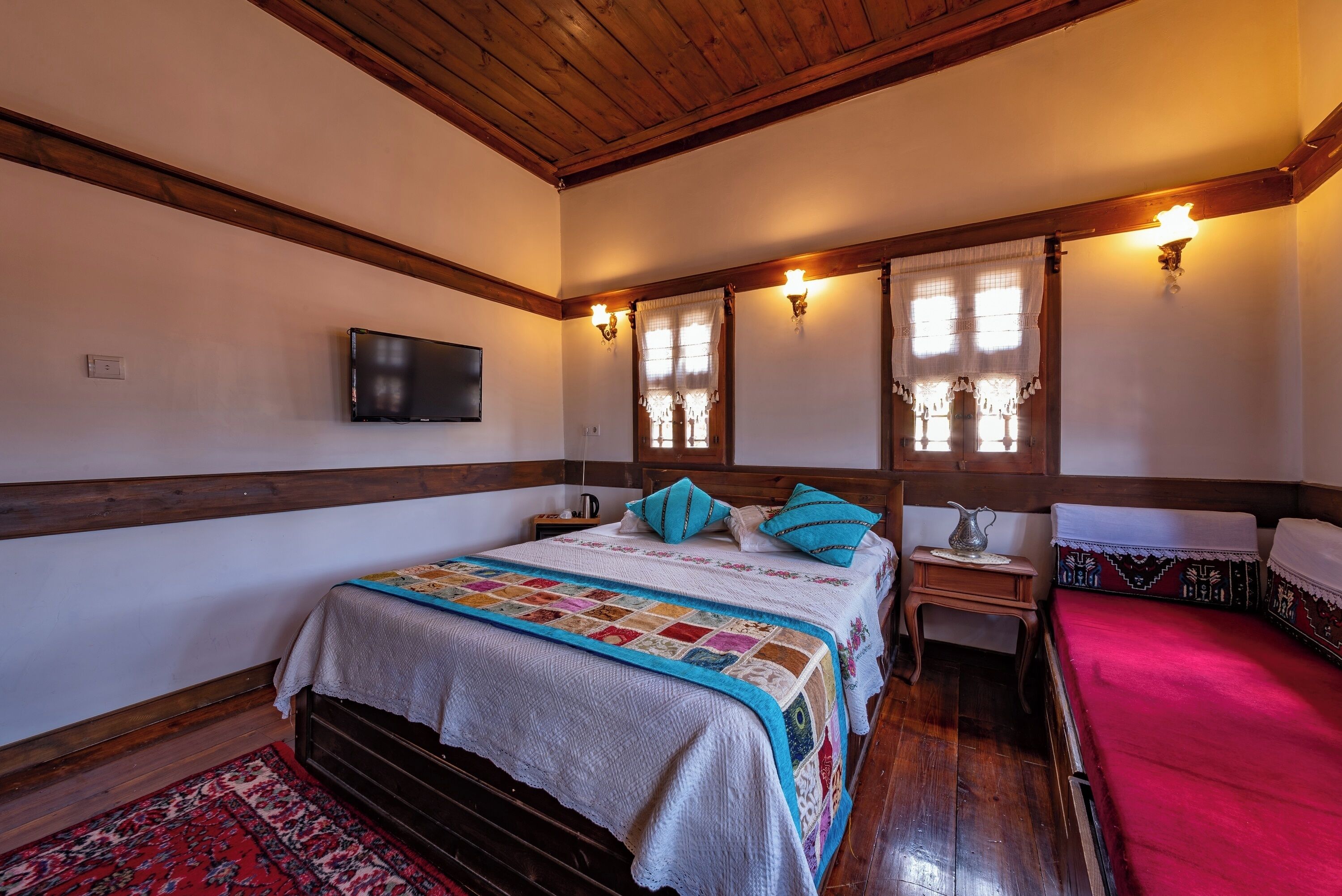 A bedroom with a wooden ceiling, a bed with a quilt, two bedside tables, a TV on the wall, and a window with curtains.