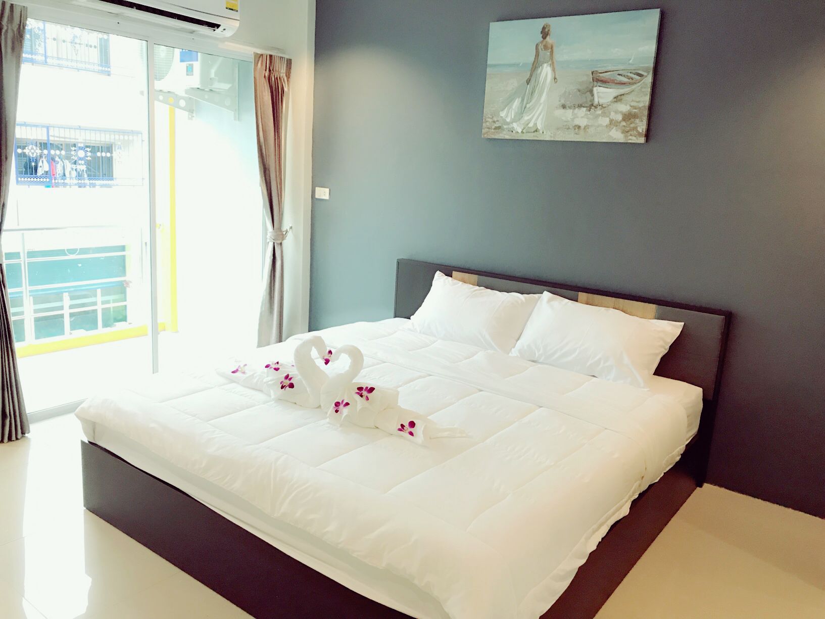Un lit bien fait, avec du linge de lit blanc et des coussins décoratifs en forme de cygne, un tableau représentant une scène de plage accroché au mur, et une vue sur un balcon avec une balustrade.