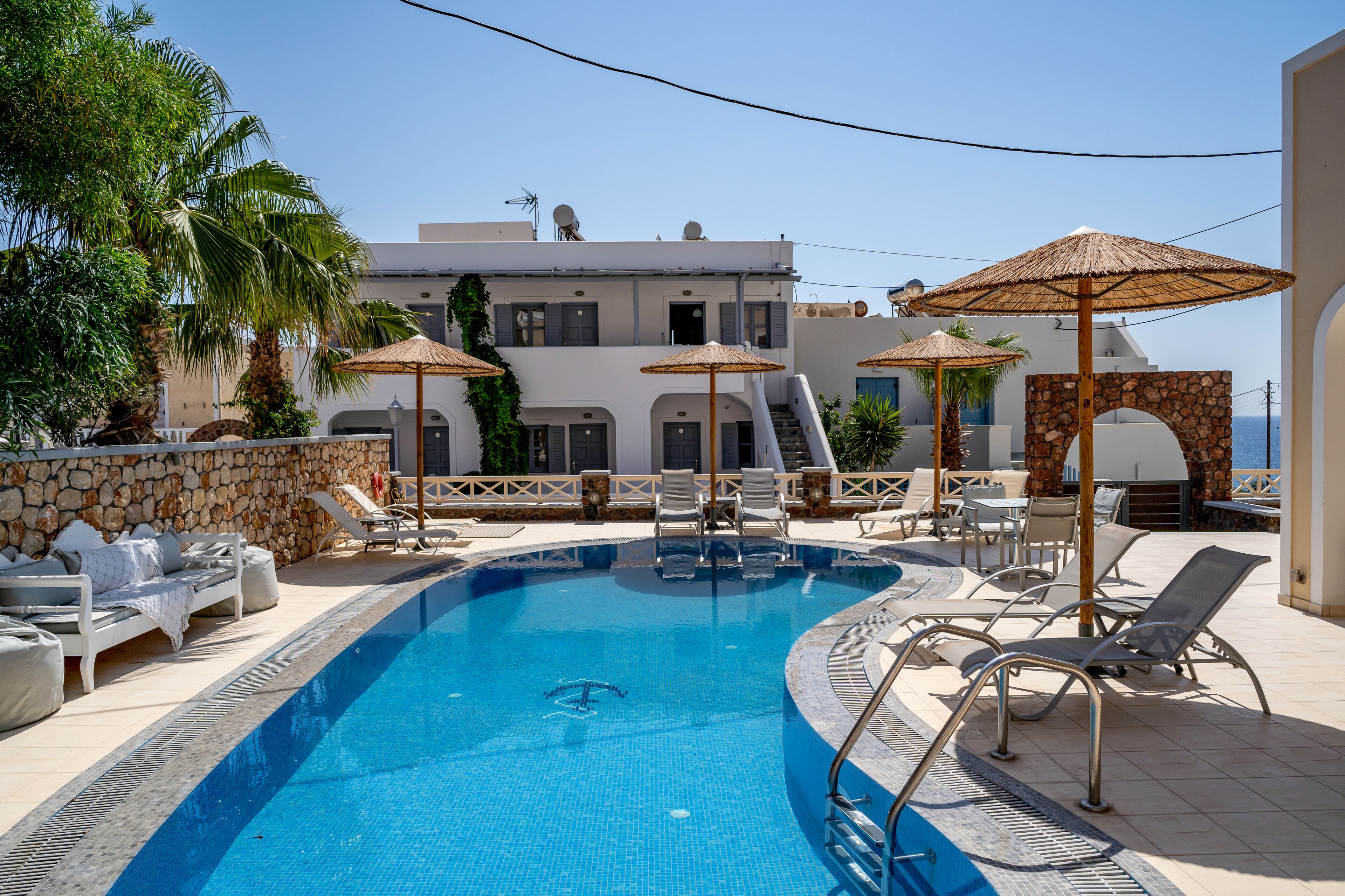 A swimming pool with lounge chairs and umbrellas, a stone wall, and a building with a white facade and blue shutters in the background.