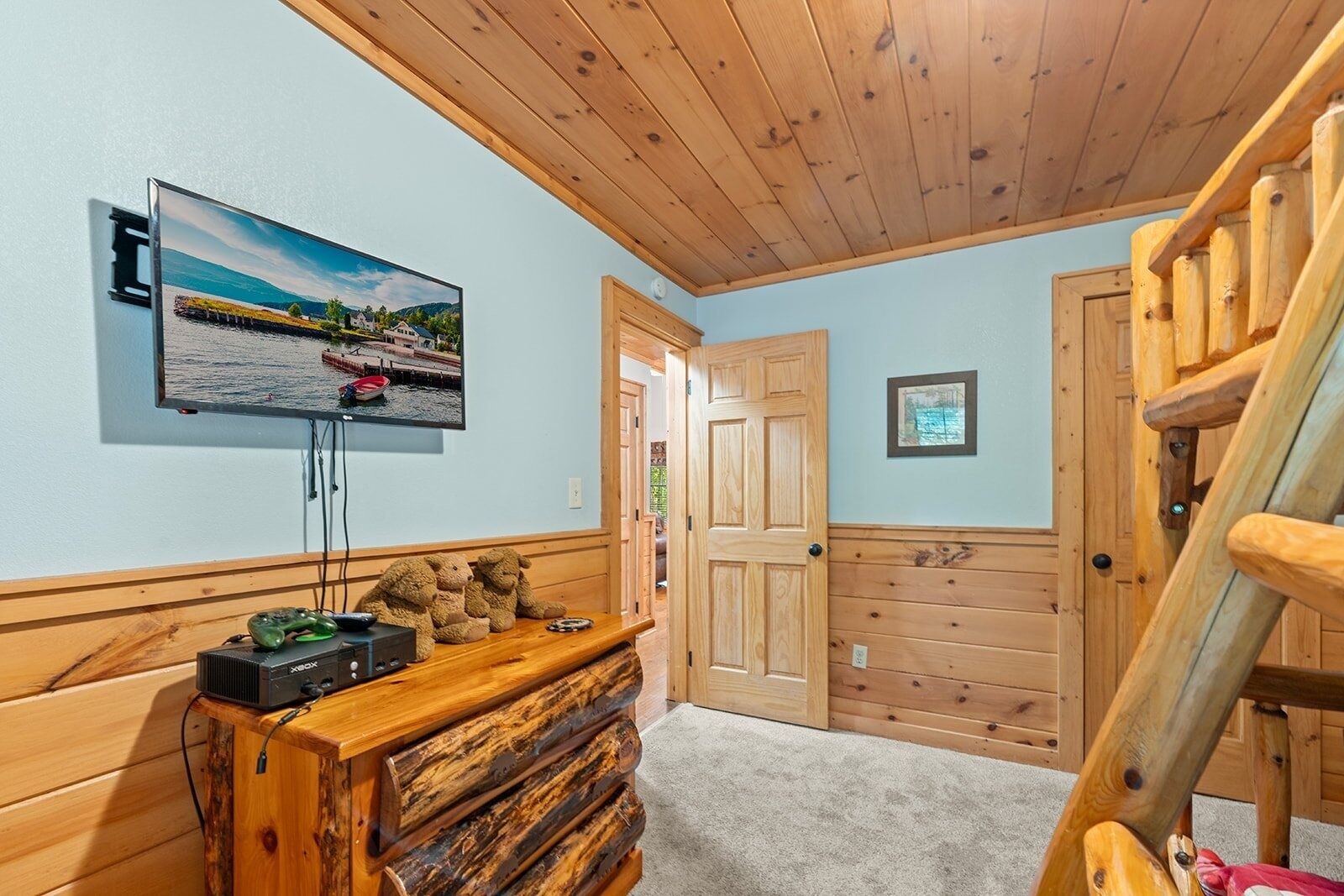 A cozy room with a wooden ceiling, a mounted TV displaying a scenic landscape, a wooden bench with plush toys, and a staircase.