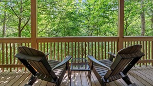 Eine Holzterrasse mit zwei Adirondack-Stühlen und einem kleinen Tisch, umgeben von einem Holzgeländer, mit Blick auf einen dichten, grünen Wald.