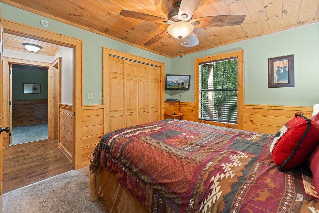A bedroom with a bed, wooden ceiling, a ceiling fan, a television, and a window with blinds.