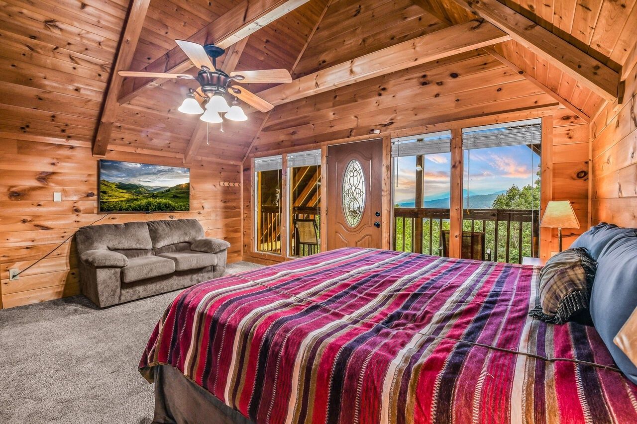 A wooden cabin bedroom with a bed, a sofa, a ceiling fan, and a large window with a view.
