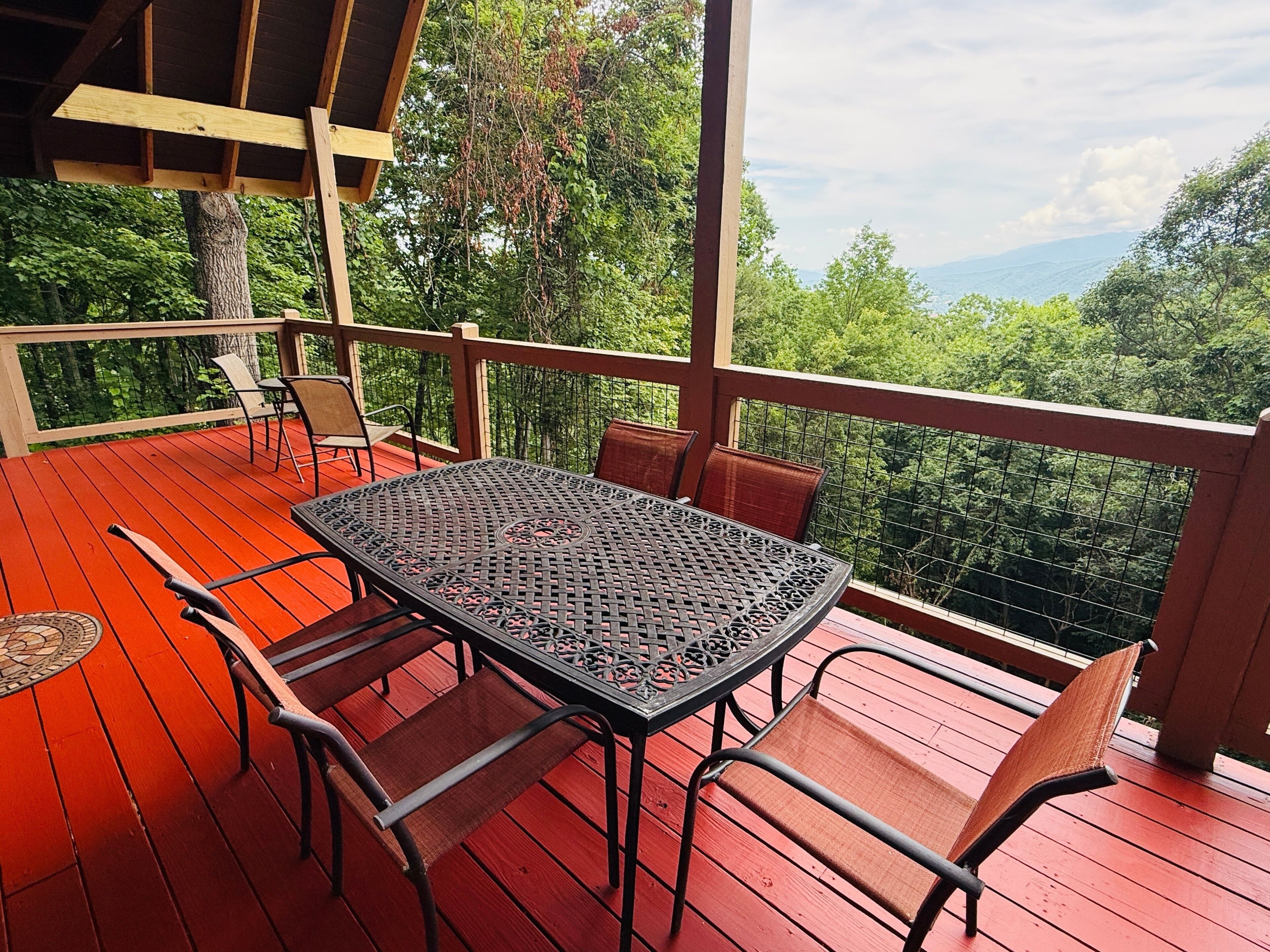 A covered outdoor deck with a table and chairs, surrounded by a wooden railing and overlooking a forested area.