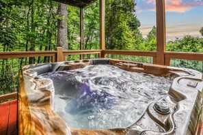A hot tub with a waterfall feature surrounded by a wooden deck and forested landscape.