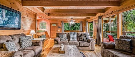A cozy cabin interior with wooden walls, a fireplace, and a large window overlooking greenery.
