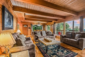 A cozy cabin interior with wooden beams, a fireplace, and a large window overlooking greenery. - Bearly Hidden Ridge 5 Bedroom Cabin by RedAwning (Gatlinburg)