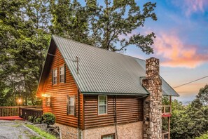 A log cabin with a stone chimney, a metal roof, and a porch with a railing.