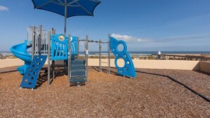 Un parque infantil con un tobogán azul, estructuras para escalar y una zona con sombra.