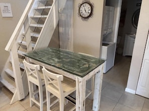 A room with a glass-top table, white chairs, a clock on the wall, and a staircase. - Seaside S 4302 1 Bedroom Condo by RedAwning (New Smyrna Beach)