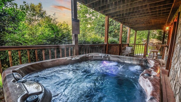 A hot tub with a wooden deck and stone walls, surrounded by lush greenery. - The Ranch Cabin on 6 Secluded Acres in the Great Smoky Mountains by RedAwning (Sevierville)