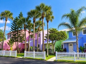 A row of colorful houses with white picket fences, palm trees, and a clear blue sky. - Coconut Palms Townhome 2 Bedroom Townhouse by RedAwning (New Smyrna Beach)