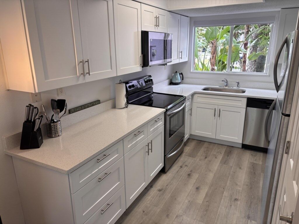 A modern kitchen with white cabinets, stainless steel appliances, and a window providing a view of greenery.
