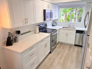 A modern kitchen with white cabinets, stainless steel appliances, and a window providing a view of greenery. - Coconut Palms Townhome 2 Bedroom Townhouse by RedAwning (New Smyrna Beach)