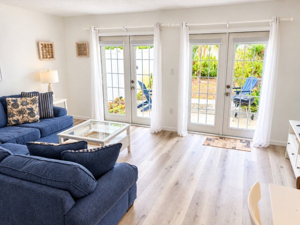 A living room with a blue sofa, a glass coffee table, and a view through glass doors to an outdoor area with chairs.