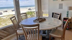 A round table with wooden chairs, a tray of decorative items, and a view of the beach and ocean through large windows.