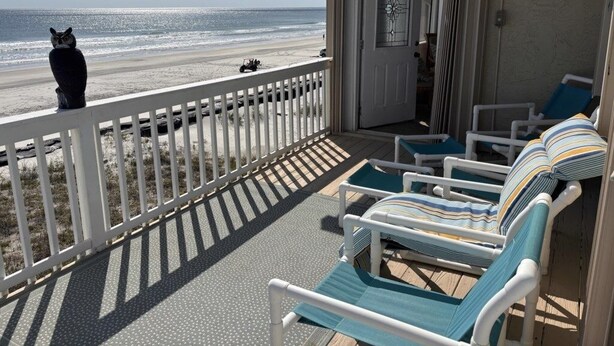 A beachside balcony with a white railing, a door with a glass panel, and two lounge chairs with striped cushions.
