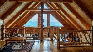 Una habitació àmplia a l'àtic amb un alt sostre de fusta, grans finestres que donen vistes a la muntanya i una barana de fusta rústica.