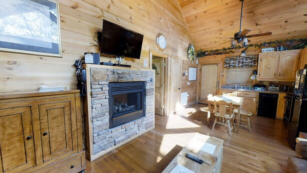 Interior de una cabaña de madera con chimenea de piedra, mesa de comedor de madera y televisor montado en la pared.