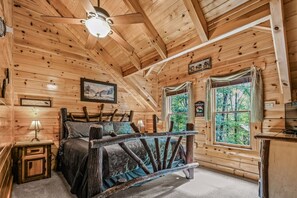 Ein Schlafzimmer in einer Blockhütte mit einem Bett, einem Nachttisch, einem Deckenventilator und einem Fenster mit Vorhängen.
