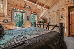 A wooden cabin bedroom with a bed, two windows, and a view of trees.
