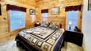 A cozy wooden cabin bedroom with a large bed, two windows with curtains, a ceiling fan, and a small bedside table with a lamp. - Picture Perfect 2 Bedroom Cabin by RedAwning (Sevierville)