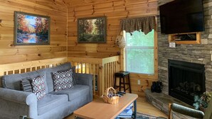 A cozy cabin interior with a wooden ceiling, a gray sofa with patterned cushions, a wooden table, a stone fireplace, and two paintings on the walls. - Picture Perfect 2 Bedroom Cabin by RedAwning (Sevierville)