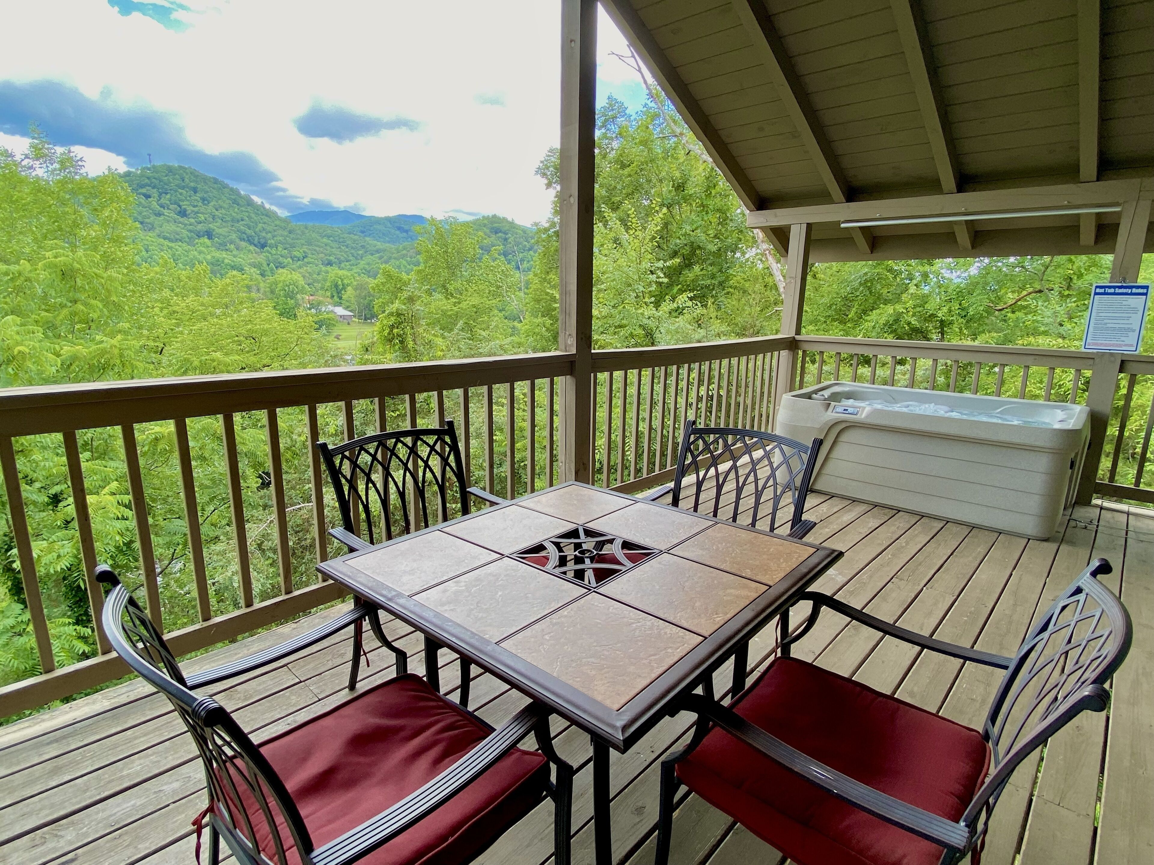 A covered outdoor deck with a table and chairs, a hot tub, and a scenic view of a forested area.