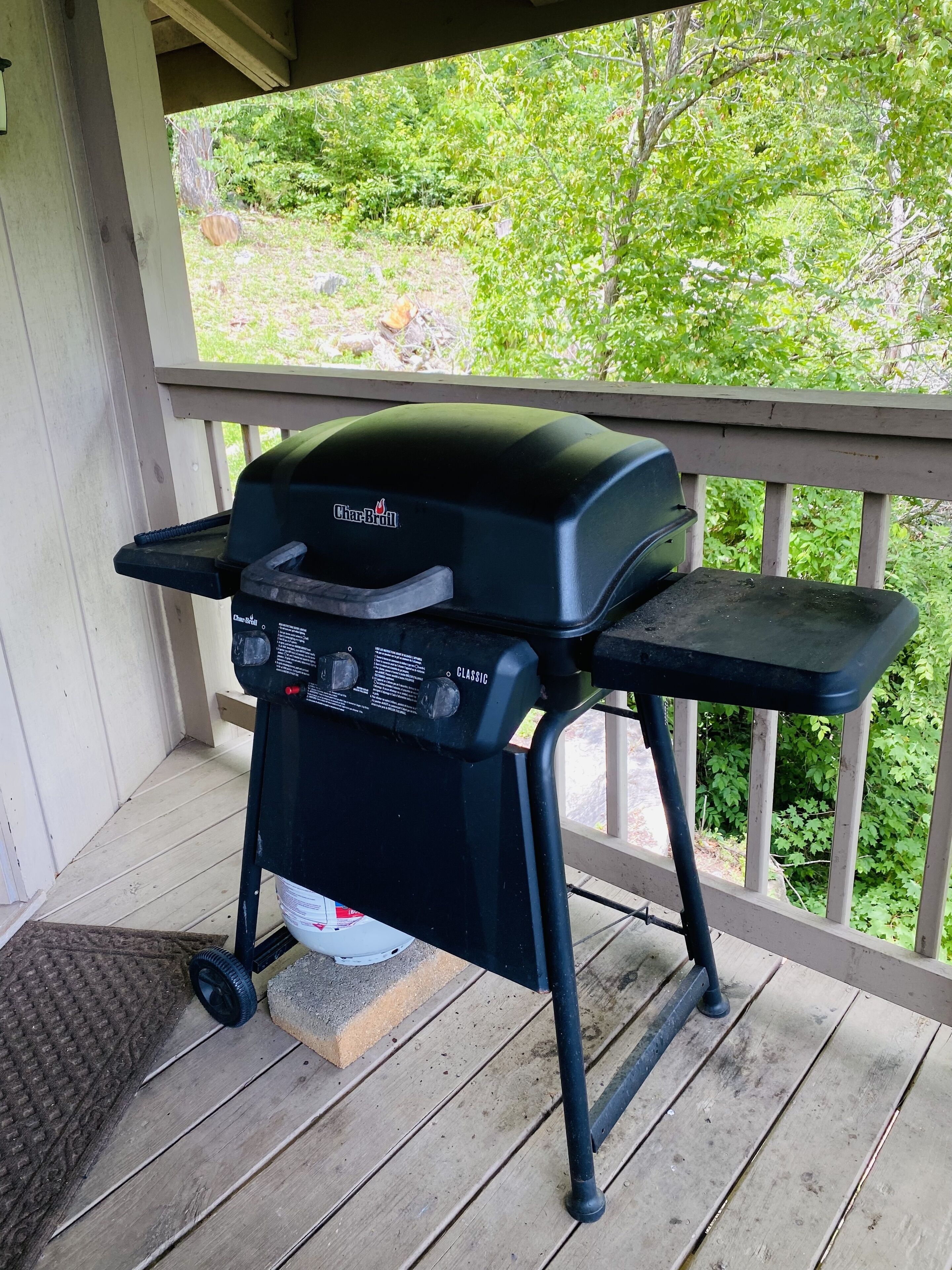 Un barbecue Char-Broil sur une terrasse en bois avec une rambarde, un paillasson et de la verdure en arrière-plan.