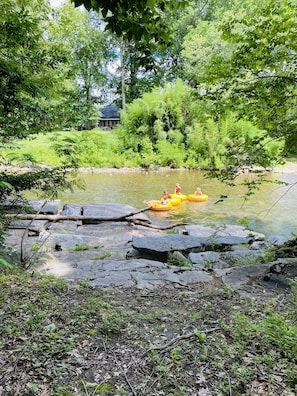 A river with rocks and greenery, a yellow raft with people, and a house in the background. - Riverview 1 Bedroom Cabin by RedAwning (Townsend)