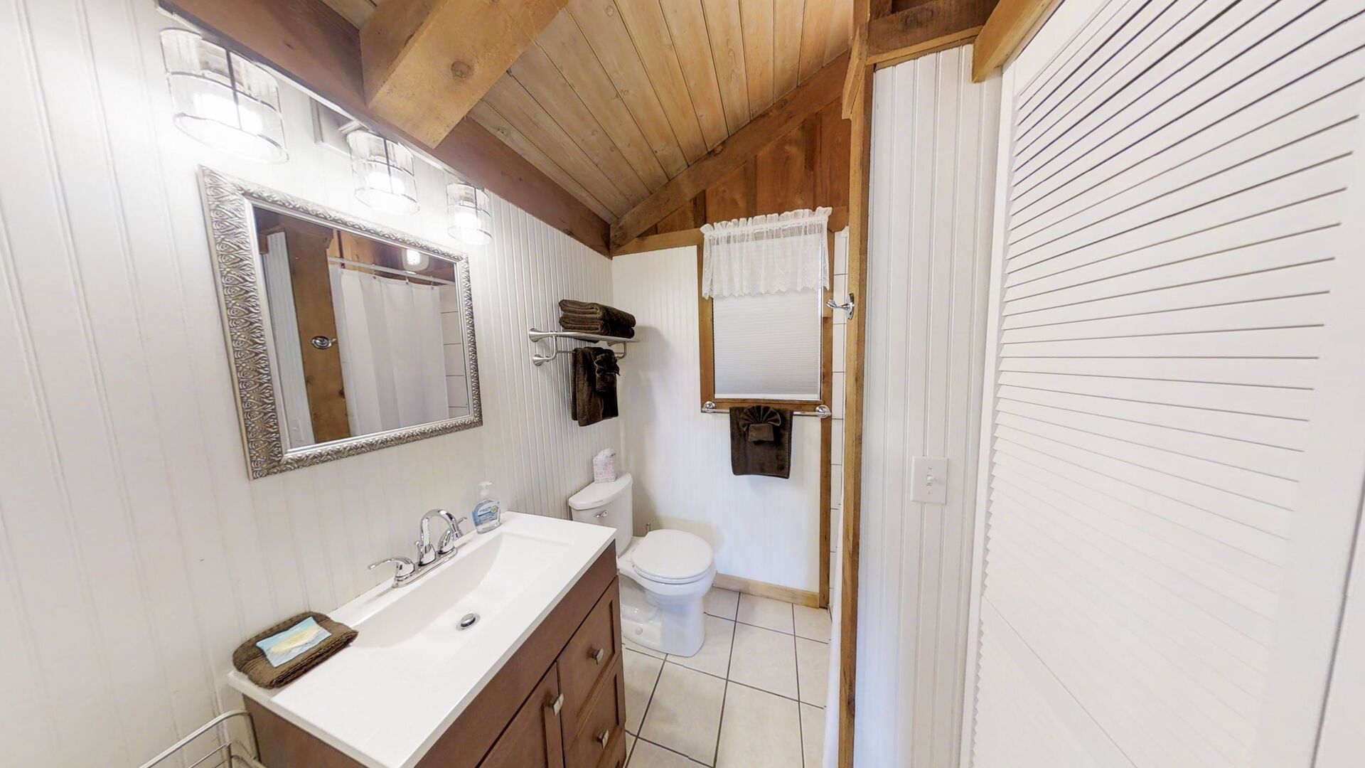 Une salle de bain avec un lavabo blanc, un miroir et des toilettes. On y trouve un porte-serviettes avec des serviettes et une fenêtre avec un rideau.