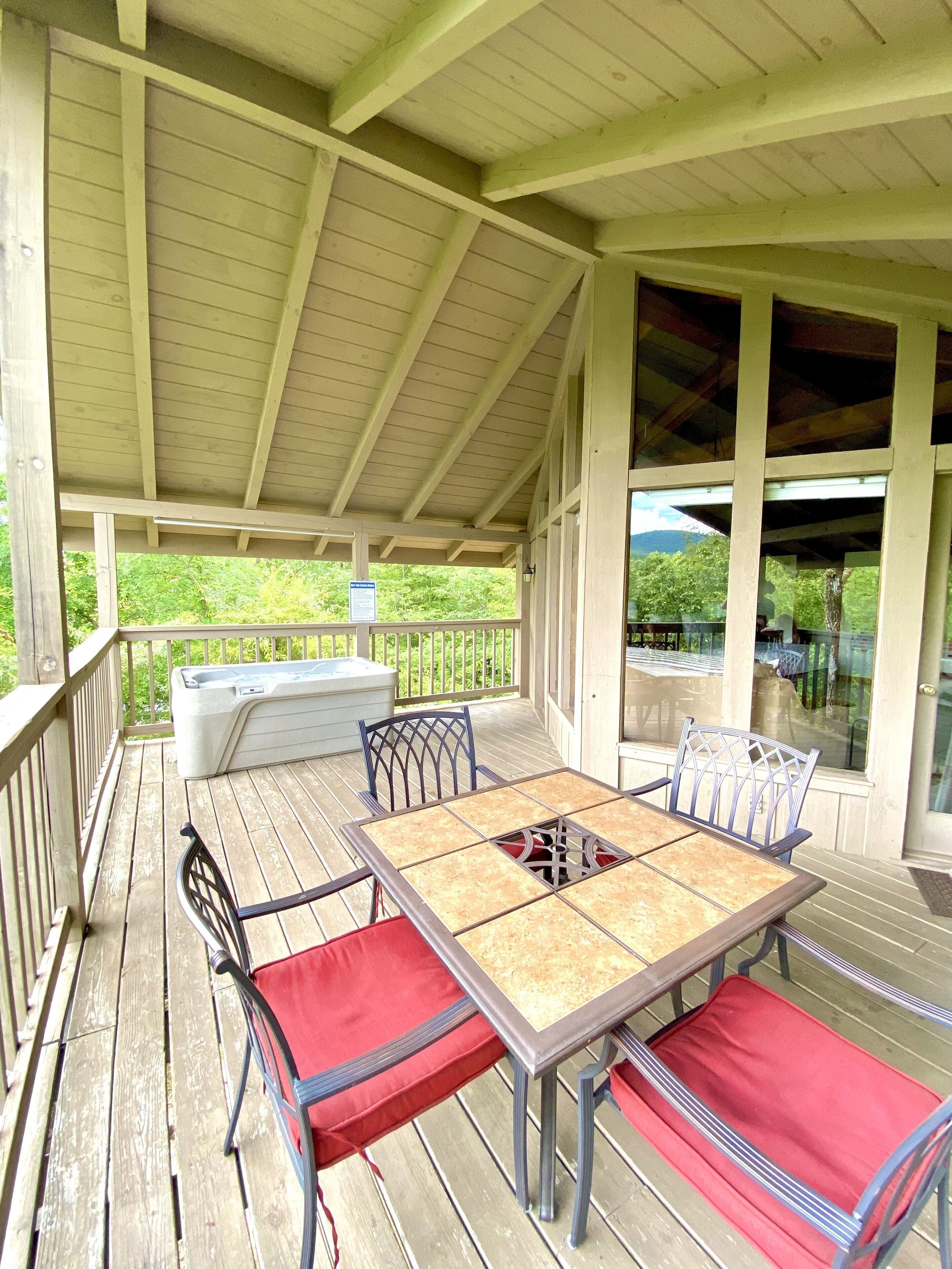 A covered outdoor deck with a wooden table and chairs, a hot tub, and a view of greenery.