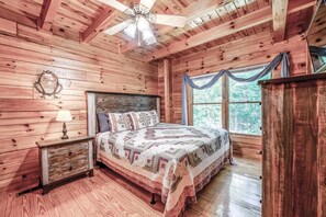Une chambre à coucher dans une cabane en bois, avec un lit, une table de chevet, un ventilateur de plafond et une fenêtre avec des rideaux.