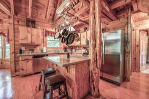 Une cuisine rustique aménagée dans une cabane en bois, avec un îlot central, des ustensiles de cuisine suspendus, un réfrigérateur en acier inoxydable et des placards en bois.