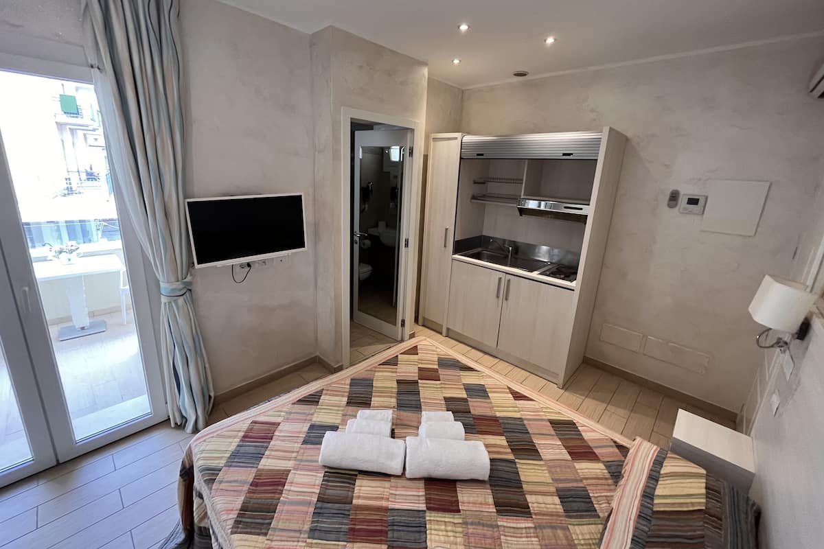A hotel room with a bed, a TV mounted on the wall, a kitchenette, and a bathroom visible through an open door.