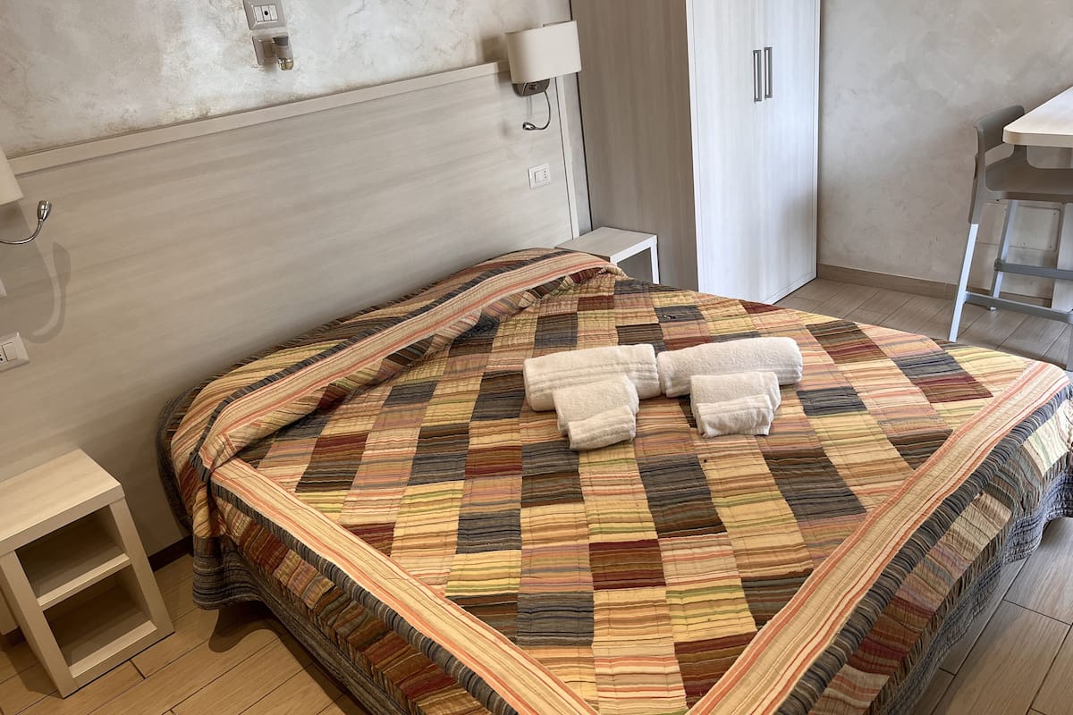 A neatly made bed with a striped quilt and two towels, a bedside table, a wall-mounted lamp, and a wardrobe in a room with a simple, clean design.