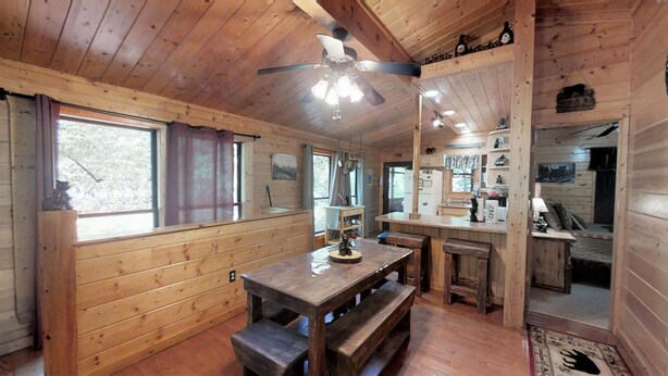 L’intérieur d’un chalet en bois comprend une table à manger, des bancs, un ventilateur de plafond et un coin cuisine visible à travers une porte ouverte.