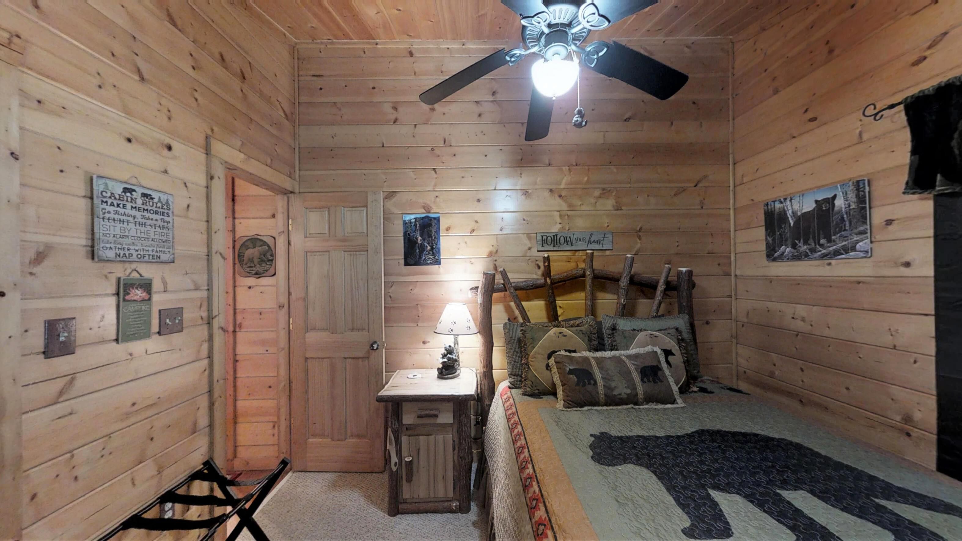 A cabin-style room with wooden walls, a ceiling fan, a bed with a bear-themed blanket, a small table with a lamp, and various wall decorations including a sign with cabin rules and a bear plaque.