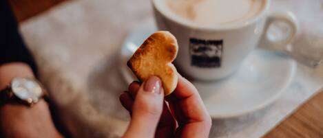 Una mano sostiene un objeto pequeño y redondo, de color marrón claro, posiblemente una galleta, frente a una taza de café con un platito blanco.