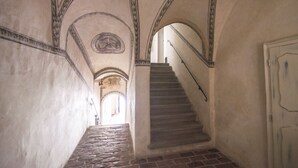 Un pasillo estrecho con una escalera de piedra que conduce a un nivel superior. Las paredes son lisas con molduras decorativas y un emblema circular en el techo.