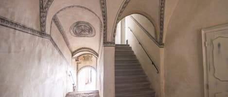 A narrow hallway with a stone staircase leading to an upper level. The walls are plain with decorative moldings and a circular emblem on the ceiling.